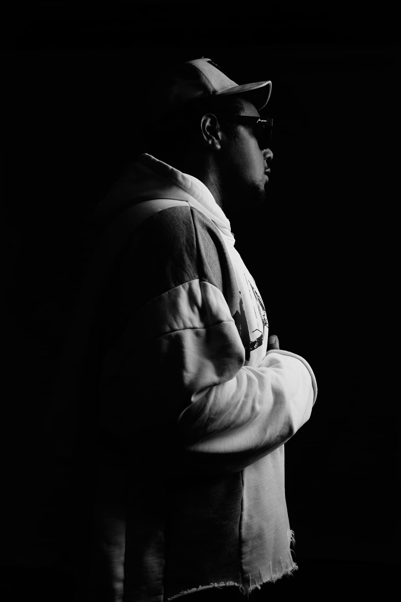 YO.G.I in profile under a single light, arms folded, in a hoodie and cap