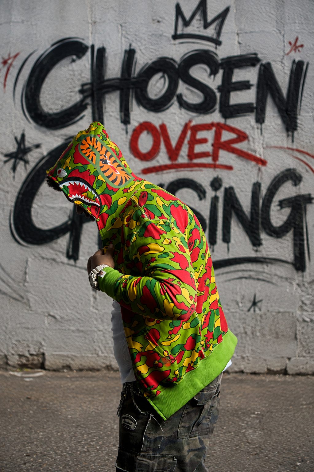 YO.G.I hooded in a bright camo shark hoodie in front of a CHOSEN OVER CHASING mural
